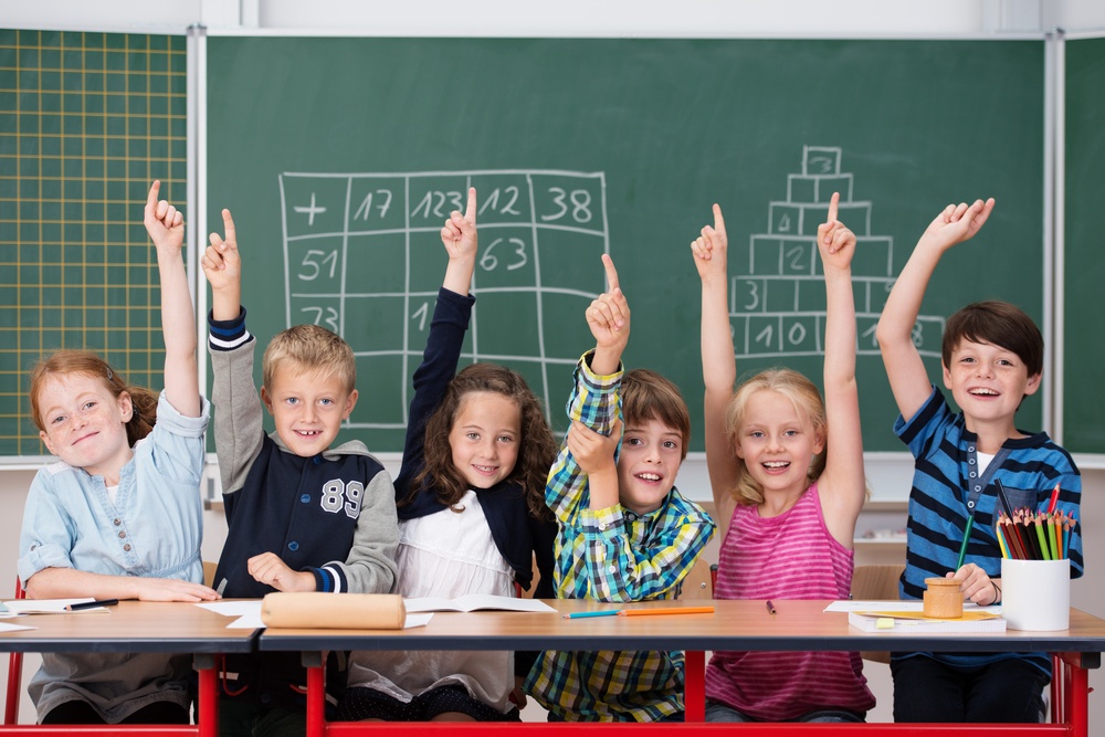 Intelligent group of young school children all raising their hands in the air to answer a question posed by the female teacher, view from behind.jpeg